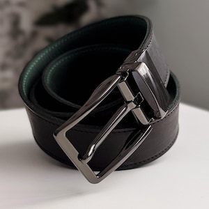 Pode incluir: Um cinto de couro preto com um design reversível. O cinto tem um interior verde escuro e uma fivela cor de metal. O cinto está enrolado sobre uma superfície branca.