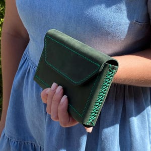 May include: A dark green leather wallet with a flap closure, held in a hand. The wallet features bright green stitching along the edges and on the flap. The stitching creates a decorative accent.