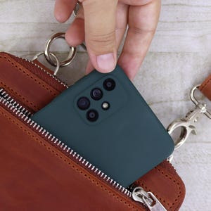 以下が含まれることがあります： 複数のカメラレンズを備えたダークティールカラーのスマートフォンが、銀色の金属製ジッパー、銀色のリングアタッチメント、銀色のスイベルクラスプ付きの茶色のレザーストラップを備えた茶色のレザー製クロスボディバッグに入れられています。