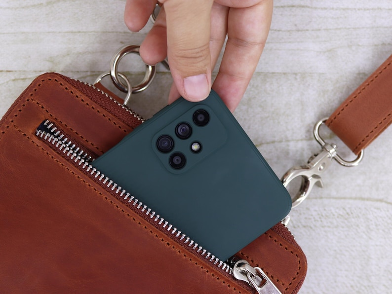 以下が含まれることがあります： 複数のカメラレンズを備えたダークティールカラーのスマートフォンが、銀色の金属製ジッパー、銀色のリングアタッチメント、銀色のスイベルクラスプ付きの茶色のレザーストラップを備えた茶色のレザー製クロスボディバッグに入れられています。