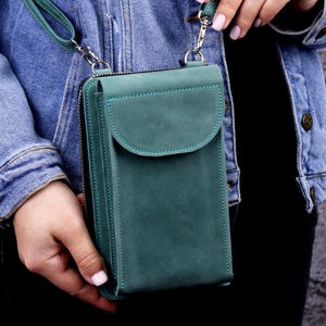 Könnte beinhalten: Türkisgrüne Leder-Umhängetasche für Telefone mit Klappenverschluss und langem Riemen. Die Tasche hat ein Reißverschlussfach und türkisfarbene Nahtdetails. Die Tasche wird von einer Person gehalten, die eine Jeansjacke trägt.