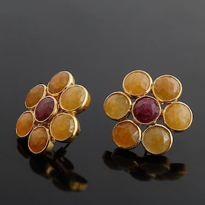 Könnte beinhalten: Ein Paar goldfarbene Ohrstecker, jeweils mit einem Cluster aus sieben Edelsteinen. Die Edelsteine sind eine Mischung aus gelben und rot-violetten Farbtönen, angeordnet in einem floralen Muster. Die Ohrringe sind auf einer reflektierenden schwarzen Oberfläche ausgestellt.