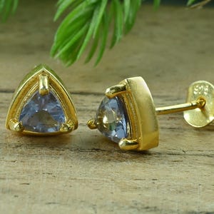 Op de afbeelding: Een paar gouden oorstekers met een driehoekige, lichtblauwe edelsteen. De oorbellen zijn gezet in een gouden bezel setting en hebben een stift en vlindersluiting. De oorbellen liggen op een houten oppervlak.
