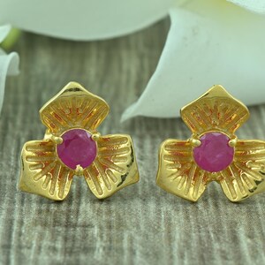 May include: A pair of gold-colored stud earrings. Each earring features a flower-shaped design with a central, round, magenta-colored gemstone. The earrings are set against a blurred background.