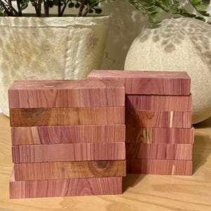May include: Two stacks of rectangular cedar wood blocks. The blocks are a reddish-brown color with visible wood grain. The blocks are stacked neatly, showcasing the natural variations in the wood.