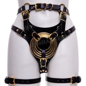 May include: Black leather harness with gold metal accents. The harness features a large circular design with multiple rings and straps. The harness is worn on a mannequin.