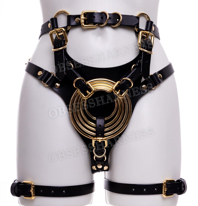 May include: Black leather harness with gold metal accents. The harness features a large circular design with multiple rings and straps. The harness is worn on a mannequin.