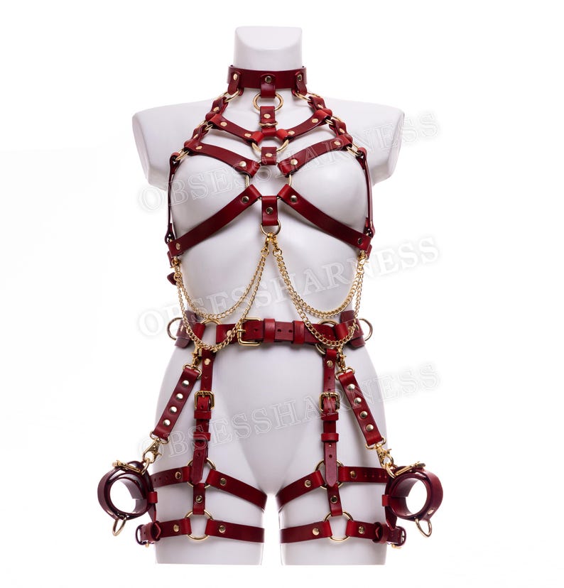 May include: A red leather harness with gold hardware and chains. The harness includes a choker, a chest piece, a belt, and thigh straps. The harness is designed with multiple straps and rings.