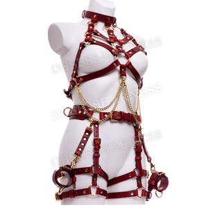 May include: A red leather harness with gold hardware and chains. The harness includes a choker, a bra, a belt, and thigh straps. The harness is designed to be worn on the body.