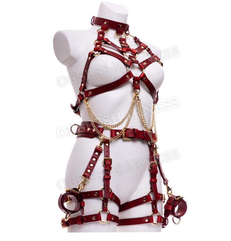 May include: A red leather harness with gold hardware and chains. The harness includes a choker, a bra, a belt, and thigh straps. The harness is designed to be worn on the body.