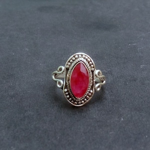 May include: A silver ring with an oval, deep red gemstone. The ring has a detailed, beaded border around the stone and decorative flourishes on the band. The ring is set against a dark gray background.