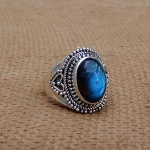 Puede incluir: Un anillo de plata con una piedra ovalada de labradorita azul engastada en un engaste de plata detallado. El anillo tiene un diseño decorativo con pequeños corazones en la banda.