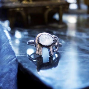 May include: A silver-toned ring in the shape of an elephant, with a round, white gemstone set in its head. The ring is displayed on a reflective surface, with the elephant's silhouette clearly visible. The ring is a piece of jewelry.
