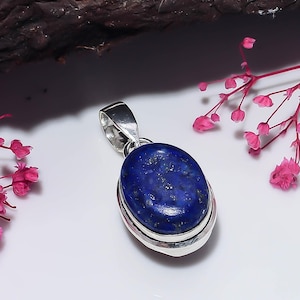 May include: An oval-shaped lapis lazuli pendant set in a polished silver frame. The deep blue stone features natural gold-colored pyrite flecks throughout its surface. A silver bail is attached to the top of the pendant for chain attachment.