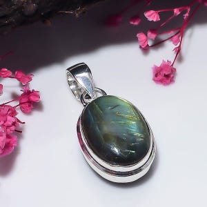 May include: An oval-shaped labradorite pendant set in a polished silver frame. The gemstone displays a dark, iridescent surface with flashes of green and gold light, featuring a smooth cabochon cut and a sturdy silver bail for attachment.