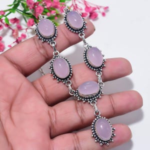 May include: A necklace featuring five oval-shaped, smooth, light pink cabochon stones set in ornate, oxidized silver-toned metal settings. The stones are linked together in a vertical arrangement, with a single pendant drop at the bottom.