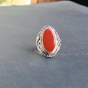 May include: A silver ring with an oval, bright red stone. The ring has a detailed, ornate design around the stone, with a beaded border and swirling patterns. The ring is set against a dark gray surface.