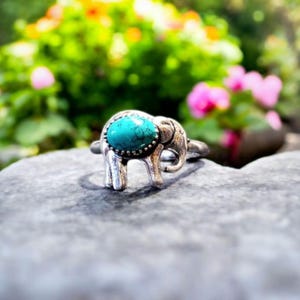 May include: A silver ring featuring an elephant design with a turquoise stone. The ring is set on a gray stone surface, with a blurred background of green foliage and pink flowers.