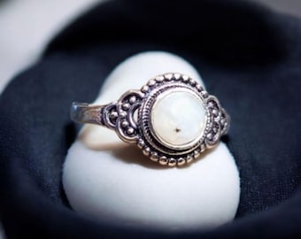 May include: A silver ring featuring a white gemstone set in an ornate silver bezel. The ring boasts a detailed design with small beads and swirls.