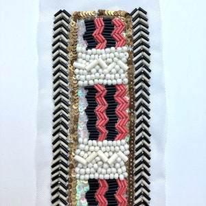 Op de afbeelding: Een rechthoekige stoffen patch met ingewikkeld kralenwerk. Het ontwerp heeft een centraal patroon van witte, zwarte en koraalkleurige kralen, omlijst door gouden pailletten en zwart-wit chevron kralen. De patch is geplaatst op een witte mesh achtergrond.
