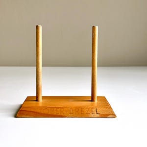 Op de afbeelding: Een houten displaystandaard met twee verticale deuvels en een rechthoekige basis. De basis is geëtst met de tekst "TOPER DREZE". De standaard is gemaakt van lichtgekleurd hout.