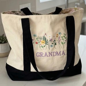 May include: A cream-colored canvas tote bag with black handles and a black base. The front features an embroidered design of colorful wildflowers and butterflies above the purple embroidered text that reads GRANDMA in capital letters.