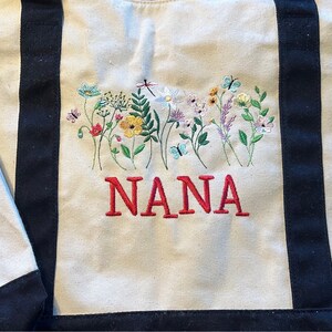 May include: A beige canvas tote bag with black vertical straps featuring an embroidered design of colorful wildflowers, butterflies, and a dragonfly. The word NANA is embroidered in red capital letters below the floral arrangement.