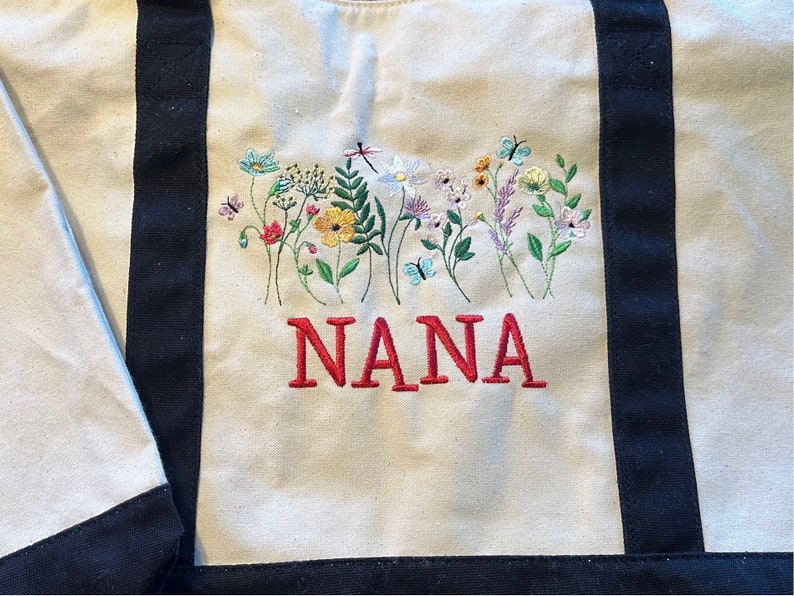 May include: A beige canvas tote bag with black vertical straps featuring an embroidered design of colorful wildflowers, butterflies, and a dragonfly. The word NANA is embroidered in red capital letters below the floral arrangement.