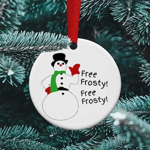 May include: White ceramic Christmas ornament with a cartoon snowman wearing a black top hat, green scarf, and red mittens. The ornament has the text "Free Frosty! Free Frosty!" printed on it.