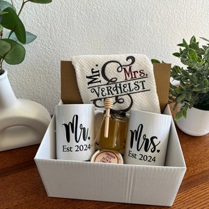May include: A white gift box filled with a wedding gift set. The set includes two white mugs with the words "Mr. Est 2024" and "Mrs. Est 2024", a white hand towel with the words "Mr. & Mrs. Verhelst" embroidered in red and black, a small jar of honey with a wooden honey dipper, and a small round container.