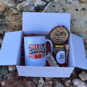 May include: A white gift box with red tissue paper inside. The box contains a white mug with a colorful sunset and the word "Colorado" on it, a small jar of honey with a label that says "Stutzman's Raw Honey Wildflower", a wooden honey dipper, and a wooden ornament with the text "The Mountains Are Calling And I Must Go".