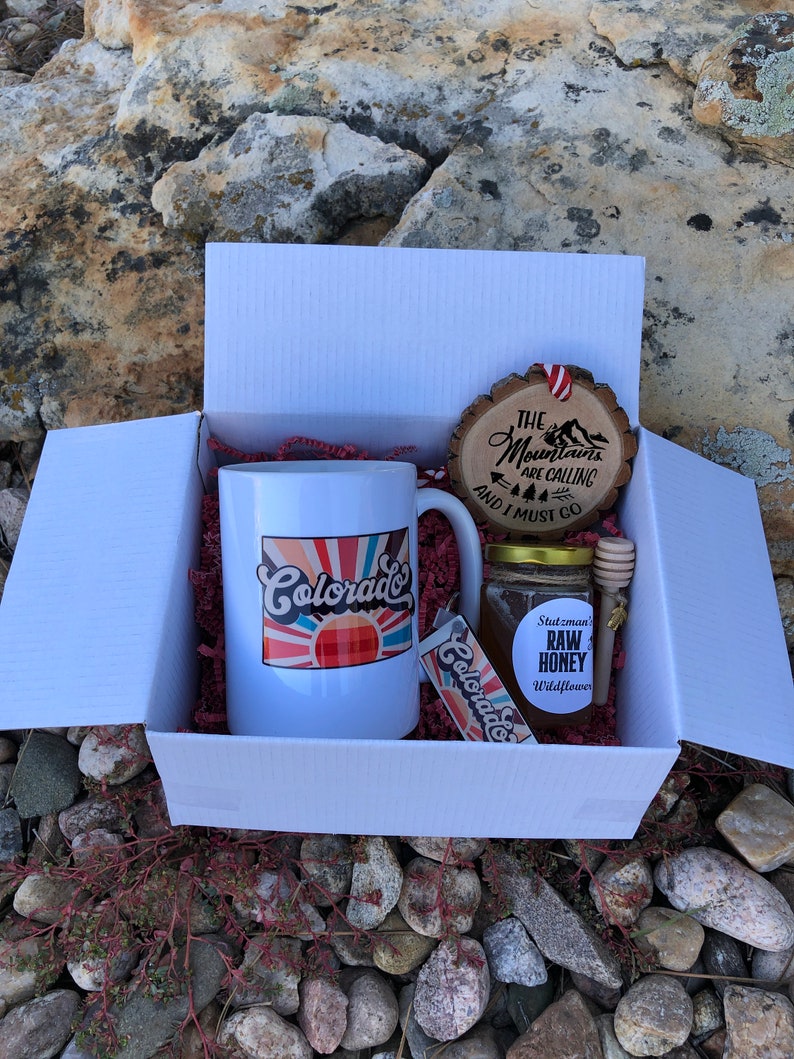 May include: A white gift box with red tissue paper inside. The box contains a white mug with a colorful sunset and the word "Colorado" on it, a small jar of honey with a label that says "Stutzman's Raw Honey Wildflower", a wooden honey dipper, and a wooden ornament with the text "The Mountains Are Calling And I Must Go".