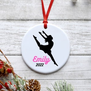 May include: White ceramic Christmas ornament with a black silhouette of a dancer in mid-air. The ornament is personalized with the name "Emily" in pink lettering and the year "2022" in black lettering.