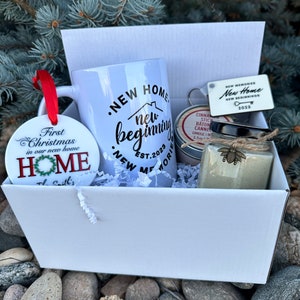 May include: A white gift box filled with a white mug with the text "New Home New Beginning Est. 2023", a small jar with a cinnamon stick candle, a small white jar with a bee design, and a white ornament with the text "First Christmas in our new home" and "New Home The Smith's".