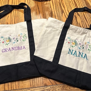 May include: Two off-white canvas tote bags with black handles and black bottom panels. Each bag features an embroidered design of colorful wildflowers and butterflies. One bag has the word GRANDMA in purple, and the other has NANA in teal.