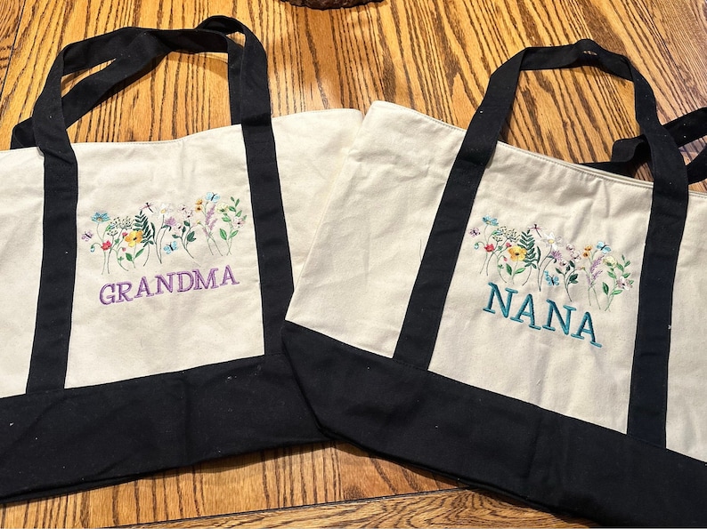 May include: Two off-white canvas tote bags with black handles and black bottom panels. Each bag features an embroidered design of colorful wildflowers and butterflies. One bag has the word GRANDMA in purple, and the other has NANA in teal.