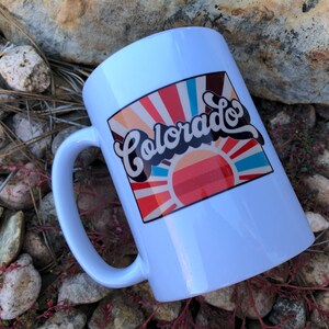 May include: White ceramic mug with a colorful graphic design featuring a sunburst and the text "Colorado" in a retro font.