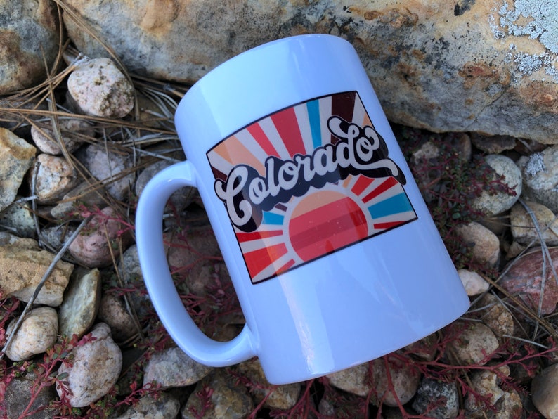 May include: White ceramic mug with a colorful graphic design featuring a sunburst and the text "Colorado" in a retro font.