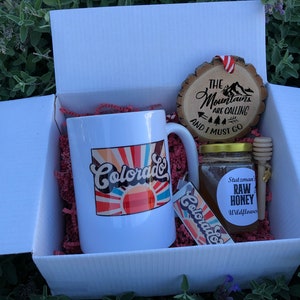 May include: A white gift box filled with a white mug with a colorful "Colorado" design, a wooden ornament with the text "The Mountains Are Calling And I Must Go", a jar of honey with a label that says "Stutzman Raw Honey Wildflower", and a wooden honey dipper.