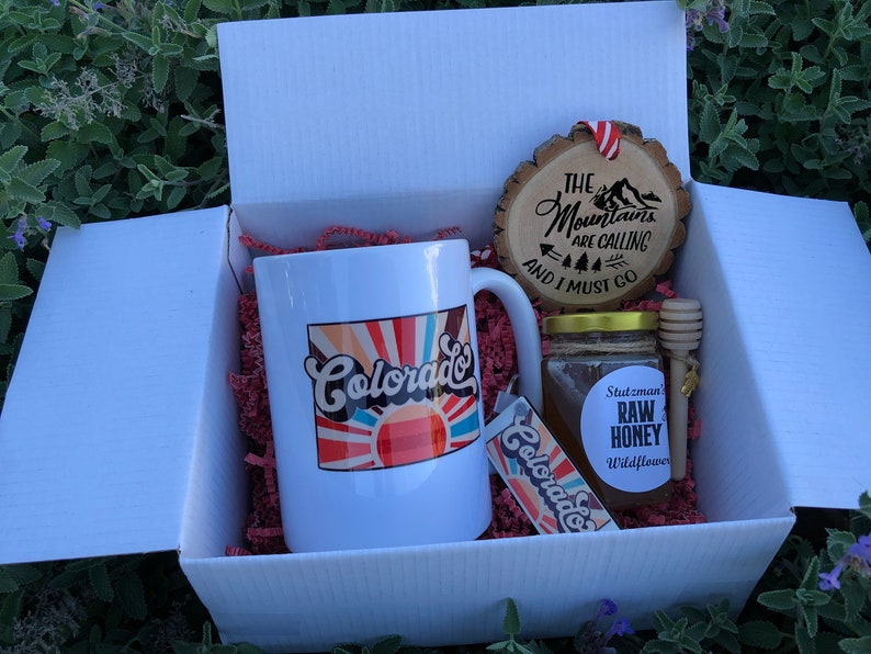 May include: A white gift box filled with a white mug with a colorful "Colorado" design, a wooden ornament with the text "The Mountains Are Calling And I Must Go", a jar of honey with a label that says "Stutzman Raw Honey Wildflower", and a wooden honey dipper.