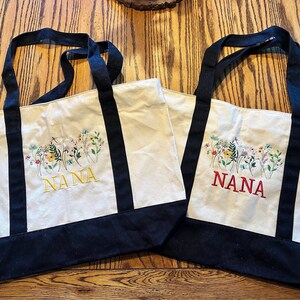 May include: Two off-white canvas tote bags with black handles and a black base. Each bag features an embroidered design of colorful wildflowers and the word NANA in capital letters, one in yellow thread and the other in red thread.