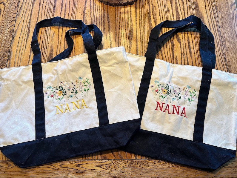 May include: Two off-white canvas tote bags with black handles and a black base. Each bag features an embroidered design of colorful wildflowers and the word NANA in capital letters, one in yellow thread and the other in red thread.