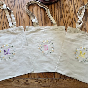 May include: Three beige canvas tote bags with embroidered floral wreaths and initials. The initials are M, D, and M. The wreaths are in shades of yellow, white, and green. The initials are in purple, pink, and yellow.