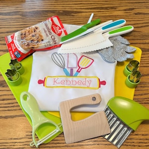 May include: A collection of baking tools and supplies, including a white apron with the name "Kennedy" embroidered on it, cookie cutters, a bag of chocolate chip cookie mix, and various kitchen utensils.