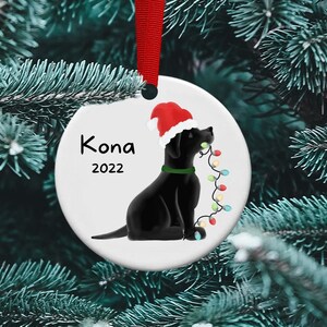 May include: A white ceramic Christmas ornament featuring a black Labrador Retriever wearing a red Santa hat and holding a string of colorful Christmas lights. The ornament is personalized with the name "Kona" and the year "2022".