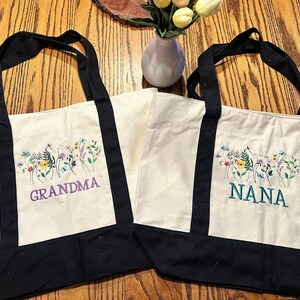 May include: Two off-white canvas tote bags with black handles and black bottom panels. Each bag features an embroidered design of colorful wildflowers and butterflies. One bag has the word GRANDMA in purple, and the other has NANA in teal.
