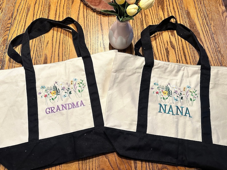 May include: Two off-white canvas tote bags with black handles and black bottom panels. Each bag features an embroidered design of colorful wildflowers and butterflies. One bag has the word GRANDMA in purple, and the other has NANA in teal.