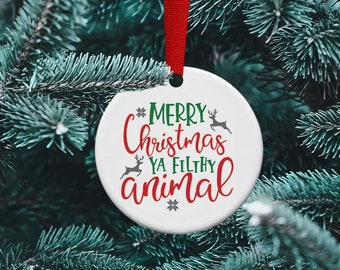 Può includere: Addobbo natalizio rotondo bianco con nastro rosso appeso a un albero di pino verde. L'addobbo ha un fiocco di neve verde e il testo "Merry Christmas Ya Filthy Animal" in lettere rosse e verdi con una grafica di renna grigia.