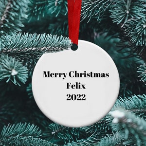 May include: White ceramic Christmas ornament with the text "Merry Christmas Felix 2022" hanging on a green pine tree branch.