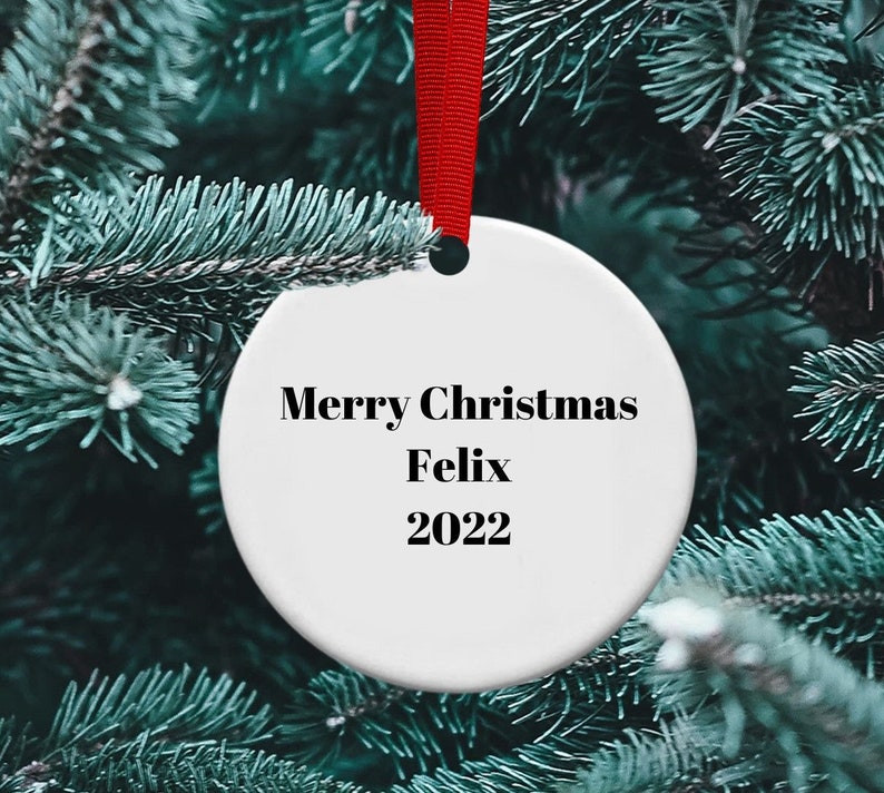 May include: White ceramic Christmas ornament with the text "Merry Christmas Felix 2022" hanging on a green pine tree branch.